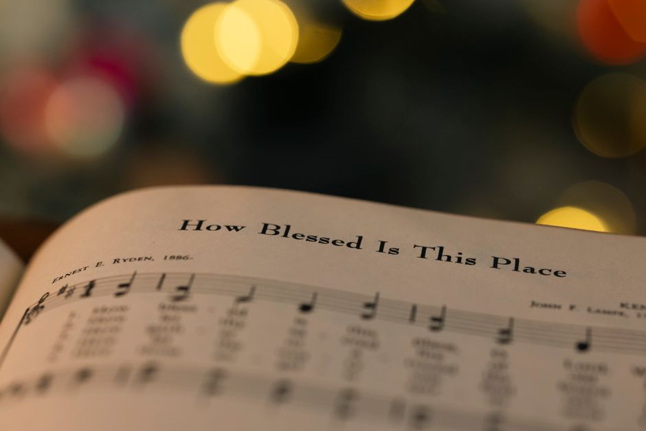 Close-up of a hymn book page with 'How Blessed Is This Place' amidst warm bokeh lights.